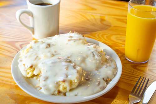 A plate of biscuits smothered in creamy gravy, accompanied by a cup of coffee and a glass of orange juice.