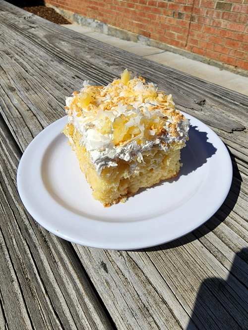 A slice of coconut cream cake topped with whipped cream and pineapple, served on a white plate on a wooden table.