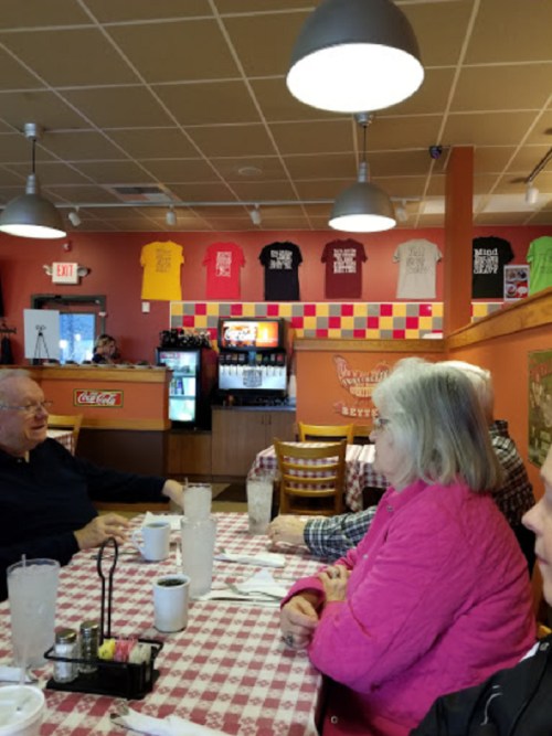 A cozy diner interior with patrons at a table, colorful shirts hanging on the wall, and a soda machine in the background.