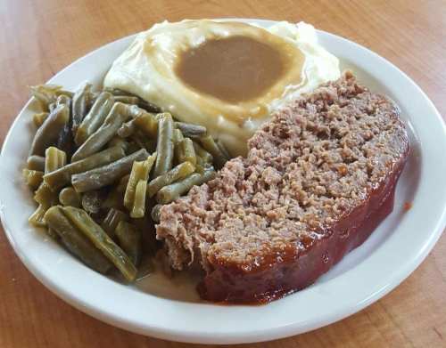 A plate of meatloaf with ketchup glaze, mashed potatoes topped with gravy, and green beans on the side.