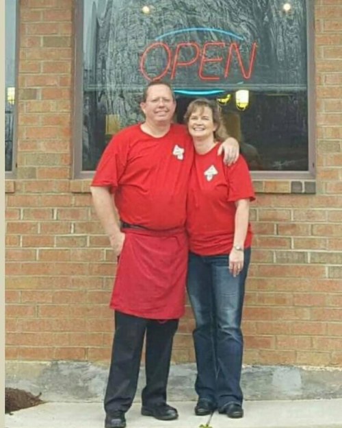 A man and woman in matching red shirts stand together outside a restaurant with an "OPEN" sign.