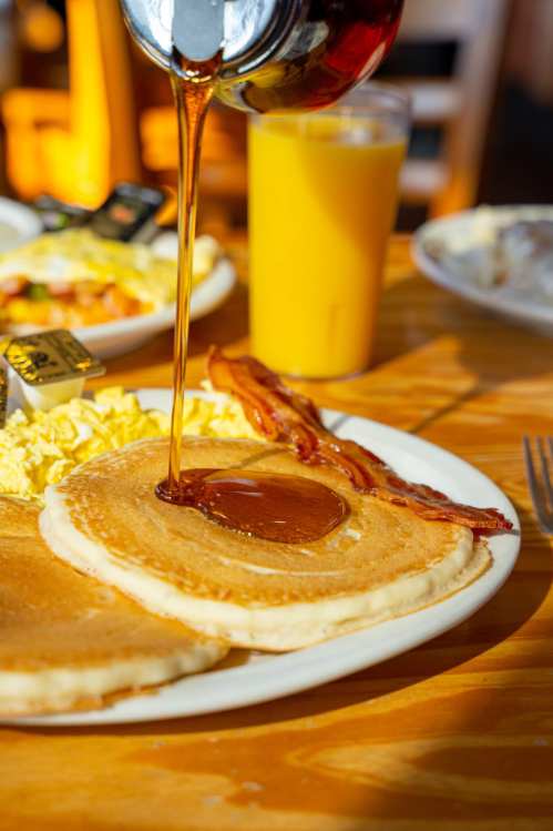 A plate of pancakes with syrup being poured, alongside bacon, scrambled eggs, and a glass of orange juice.