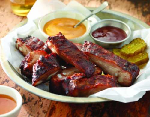 A platter of barbecue ribs with dipping sauces and pickles on a rustic wooden table.