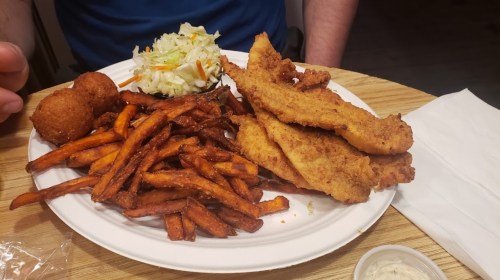A plate of fried fish fillets, sweet potato fries, coleslaw, and hushpuppies, with a dipping sauce on the side.