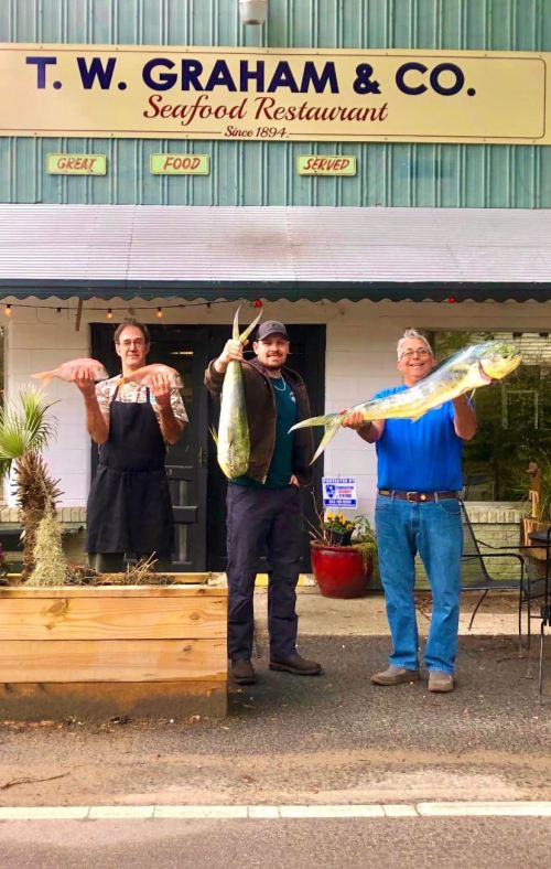 Three men stand outside T.W. Graham & Co. Seafood Restaurant, each holding a large fish, smiling at the camera.