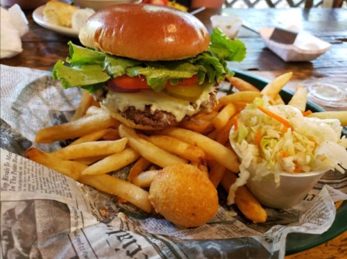 A juicy burger with lettuce, tomato, and mayo, served with fries, coleslaw, and a hushpuppy on a rustic table.