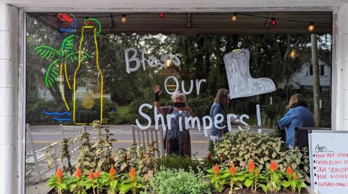A window display with "Bless Our Shrimpers!" written, featuring a neon bottle and palm tree, and people outside.