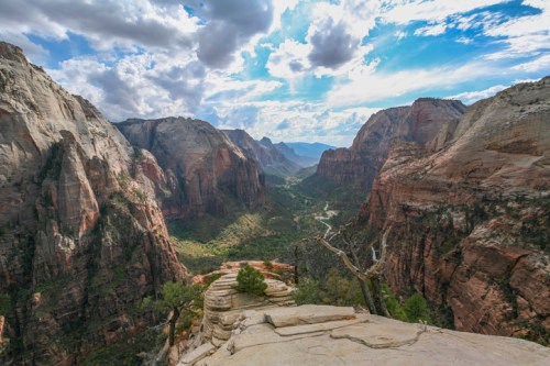 A panoramic view of Zion National Park, showcasing towering red rock cliffs and a lush green valley under a cloudy sky.
