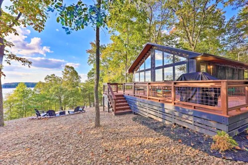 A modern cabin with large windows, surrounded by trees, overlooking a lake, with a deck and outdoor seating area.