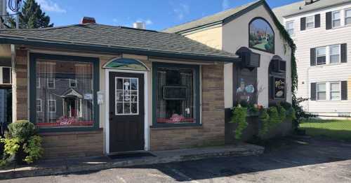 A small deli with a stone exterior, large windows, and a mural on the side, surrounded by greenery and a blue sky.