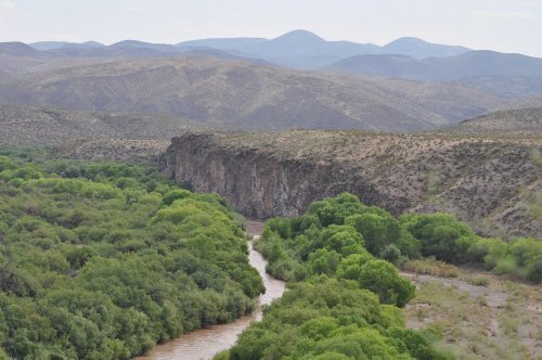 A winding river flows through lush greenery, bordered by rocky cliffs and distant mountains under a cloudy sky.