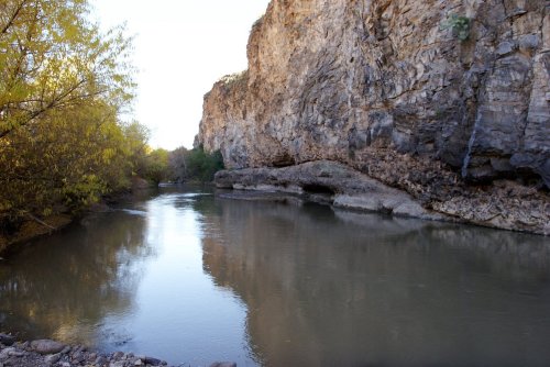 A serene river flows between rocky cliffs, surrounded by trees and reflecting the clear sky above.