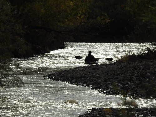 A silhouette of a person kayaking on a shimmering river surrounded by trees and rocky banks.