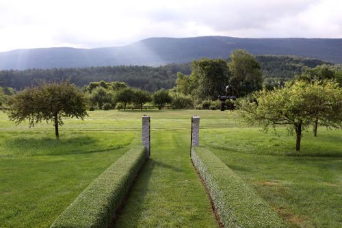 A serene landscape featuring neatly trimmed hedges leading to a lush green field and distant mountains under a cloudy sky.
