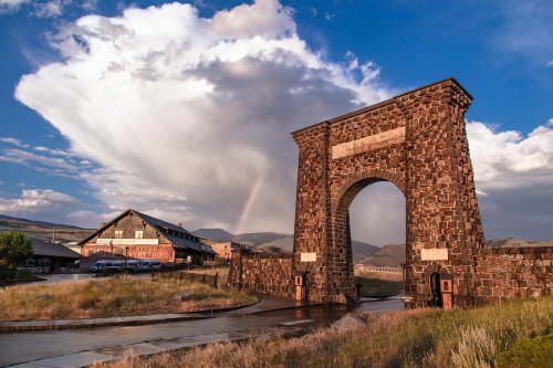 A stone archway stands against a dramatic sky with clouds and a rainbow, near a rustic building in a scenic landscape.