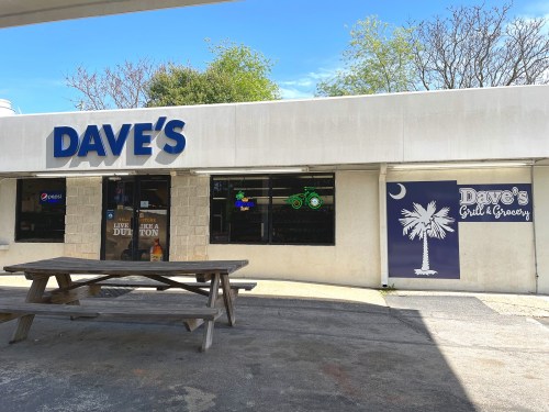 Exterior of Dave's Grill & Grocery, featuring a sign, windows with displays, and a picnic table in front.
