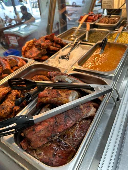 A variety of grilled meats and sides displayed in metal trays at a food counter, with tongs resting on the ribs.