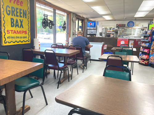 A quiet diner with empty tables, a man sitting alone, and a counter with snacks and drinks in the background.