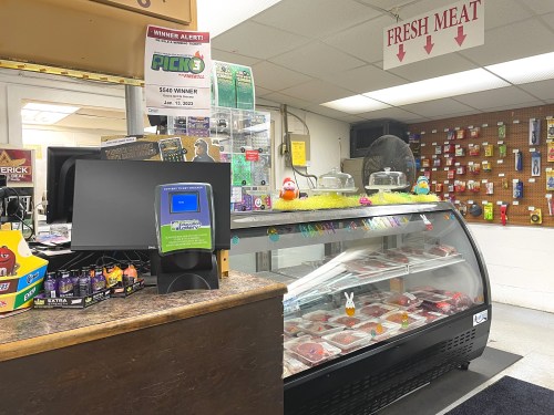 A store interior featuring a meat display case, lottery machine, and various items on a counter and wall.