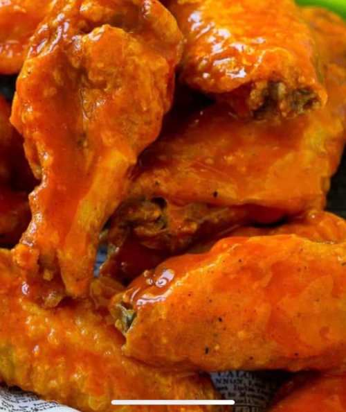 A close-up of crispy chicken wings coated in a spicy orange sauce, piled together on a plate.