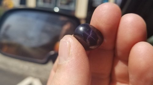 A person holding a small, dark purple gemstone with a shiny surface, with a car side mirror in the background.