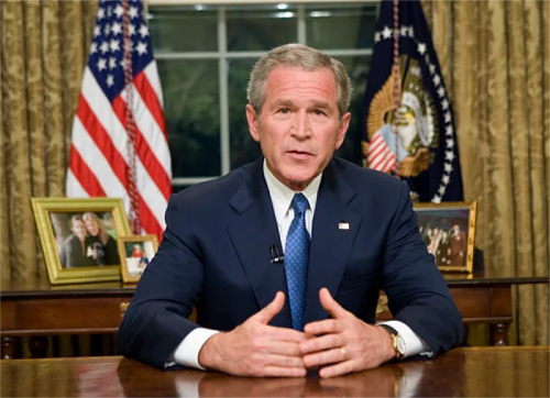 George W. Bush seated at a desk in the Oval Office, speaking with flags and family photos in the background.