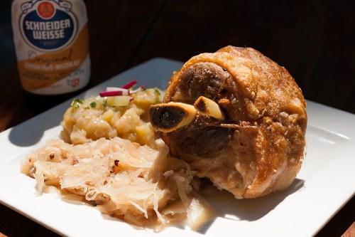 A plate featuring crispy pork knuckle, sauerkraut, potato salad, and a bottle of Schneider Weisse beer in the background.