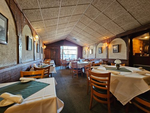 Cozy restaurant interior with tables set for dining, warm lighting, and a view of the outside through large windows.