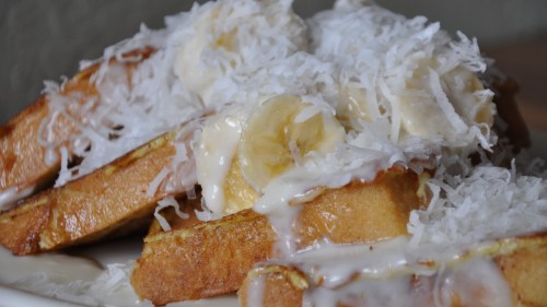 A close-up of French toast topped with banana slices, shredded coconut, and a creamy glaze.