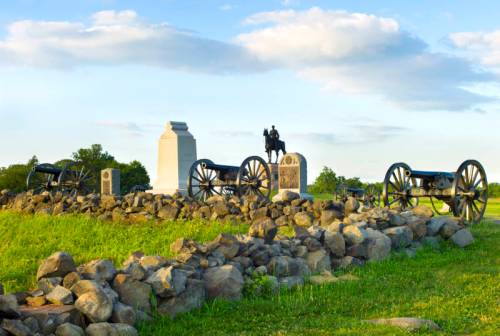 Historic battlefield scene featuring cannons, monuments, and a statue on a grassy landscape under a blue sky.