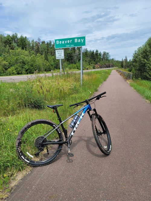 A mountain bike rests on a paved path beside a sign for Beaver Bay, surrounded by greenery and trees.