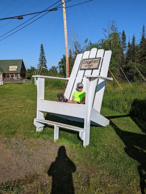 A person in a bright green shirt sits in a large white Adirondack chair, with trees and a building in the background.