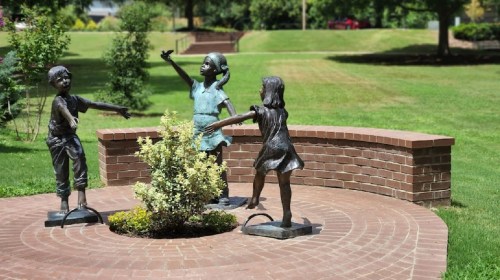 Three bronze statues of children playing in a grassy park, surrounded by a circular brick path and a small shrub.