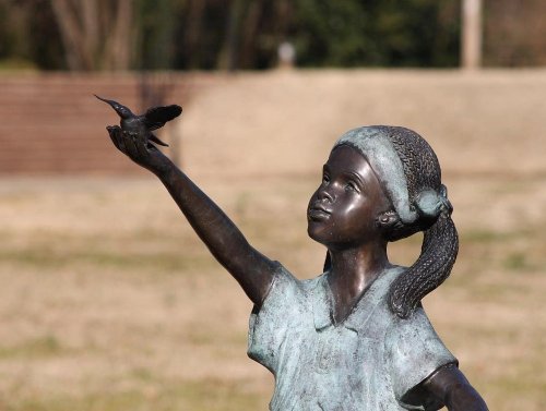 A bronze statue of a girl reaching out to a small bird, set in a grassy outdoor area.
