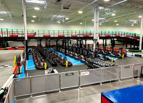 An indoor go-kart track with multiple karts lined up, surrounded by barriers and a multi-level viewing area.
