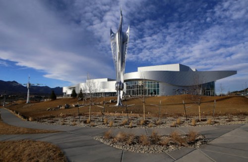 A modern building with a large silver sculpture in front, set against a backdrop of mountains and a blue sky.