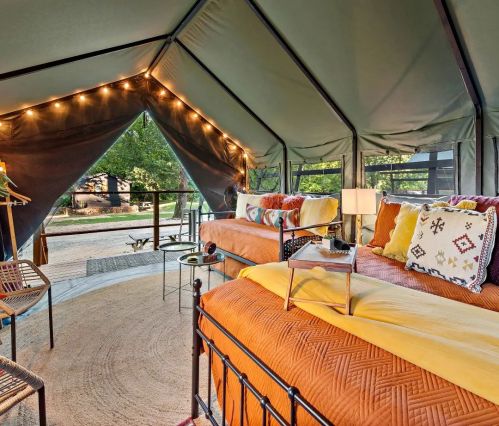Cozy glamping tent interior with two beds, colorful pillows, and warm lighting, surrounded by nature.