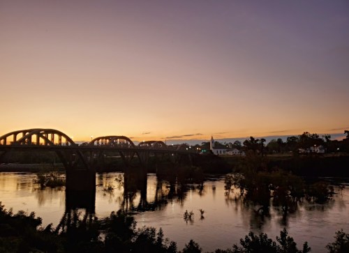 A serene sunset over a river, featuring a bridge and silhouettes of trees along the water's edge.