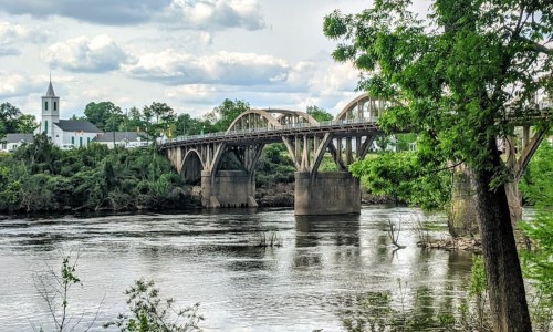 A bridge spans a river, surrounded by greenery and a church in the background under a cloudy sky.
