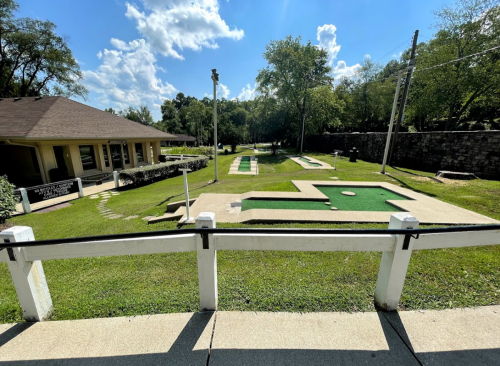 A mini-golf course with green turf, surrounded by trees and a building in the background under a blue sky.