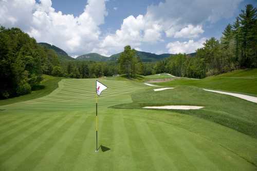 A scenic golf course with a flag on the green, surrounded by lush trees and mountains under a partly cloudy sky.