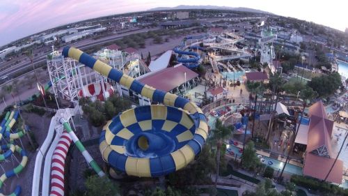 Aerial view of a colorful water park featuring slides, pools, and attractions surrounded by palm trees and buildings.