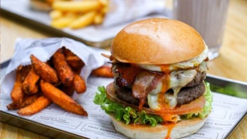 A juicy burger with lettuce, tomato, cheese, and bacon, served with sweet potato fries and a milkshake in the background.