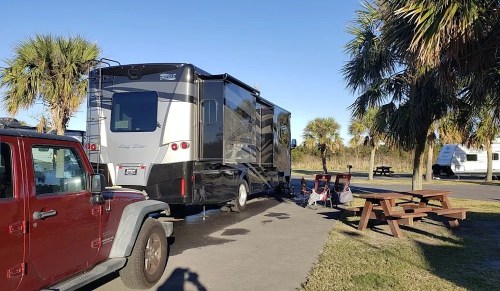 A black RV parked beside palm trees, with a red Jeep and picnic tables in a sunny outdoor setting.