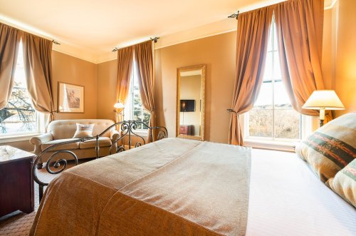 Cozy hotel room with a bed, sofa, large windows, and warm-toned decor. Natural light fills the space.