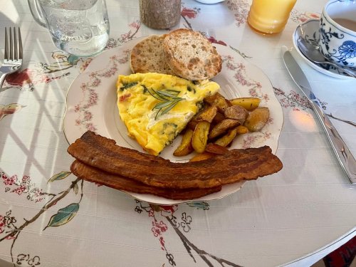 A plate with an omelette, roasted potatoes, crispy bacon, and two slices of bread, set on a floral tablecloth.