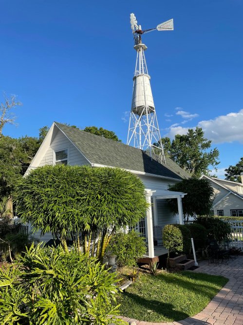A charming white house with a windmill in the background, surrounded by lush greenery and a clear blue sky.