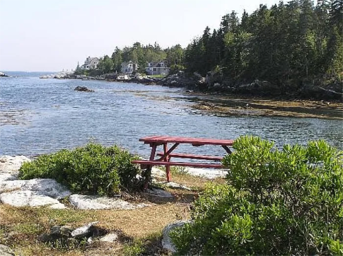 A red picnic table by a calm coastal scene with trees and houses in the background.