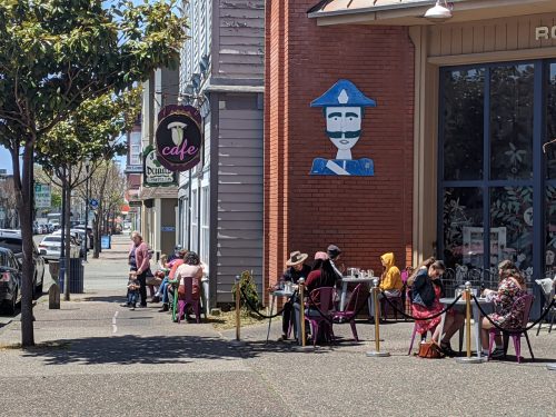 Outdoor café scene with people dining at tables, a mural of a police officer on the wall, and trees lining the street.