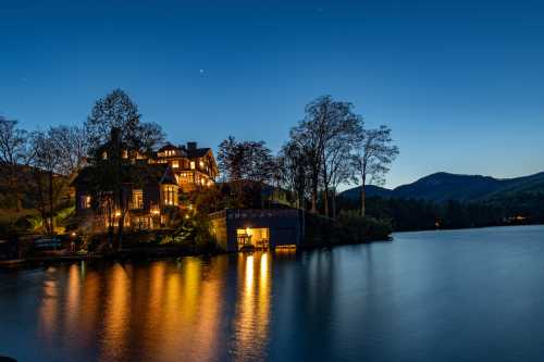 A serene lakeside house illuminated at dusk, reflecting on calm waters with mountains in the background.